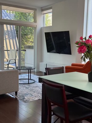 Ein modernes Wohnzimmer mit Esstisch, Stühlen, einem an der Wand befestigten Fernseher und einem großen Fenster mit Blick ins Freie.