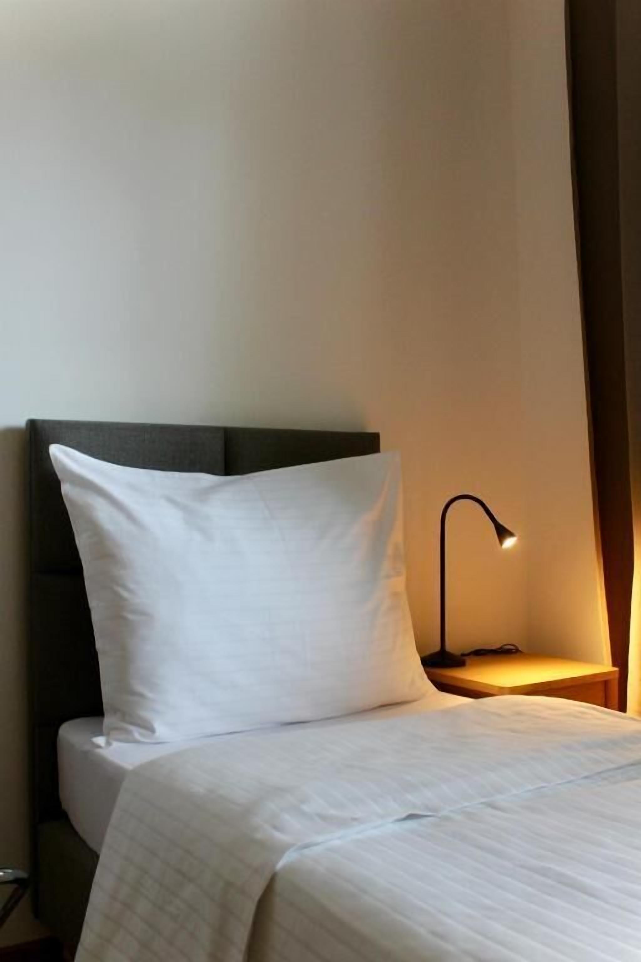 A neatly made bed with a white pillow and a dark headboard, a bedside lamp, and a plain wall.