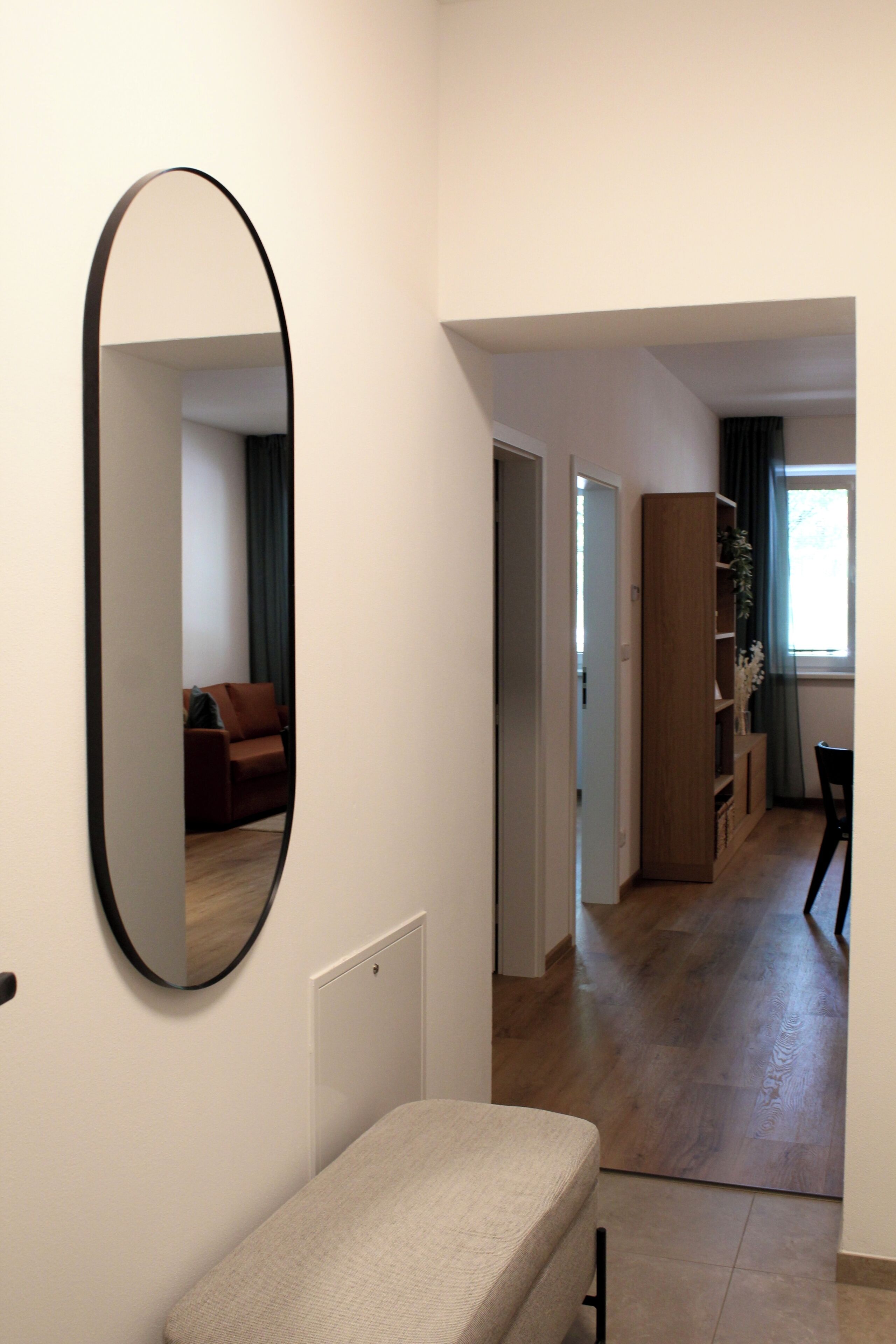 A hallway with a bench, a mirror, and a door leading to a living area with a sofa and a plant.