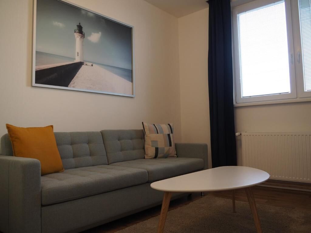 Uma sala de estar com um sofá cinza, uma mesa de centro branca, uma pintura de um farol e uma janela com cortinas.
