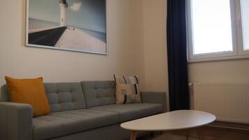 Uma sala de estar com um sofá cinza, uma mesa de centro branca, uma pintura de um farol e uma janela com cortinas.
