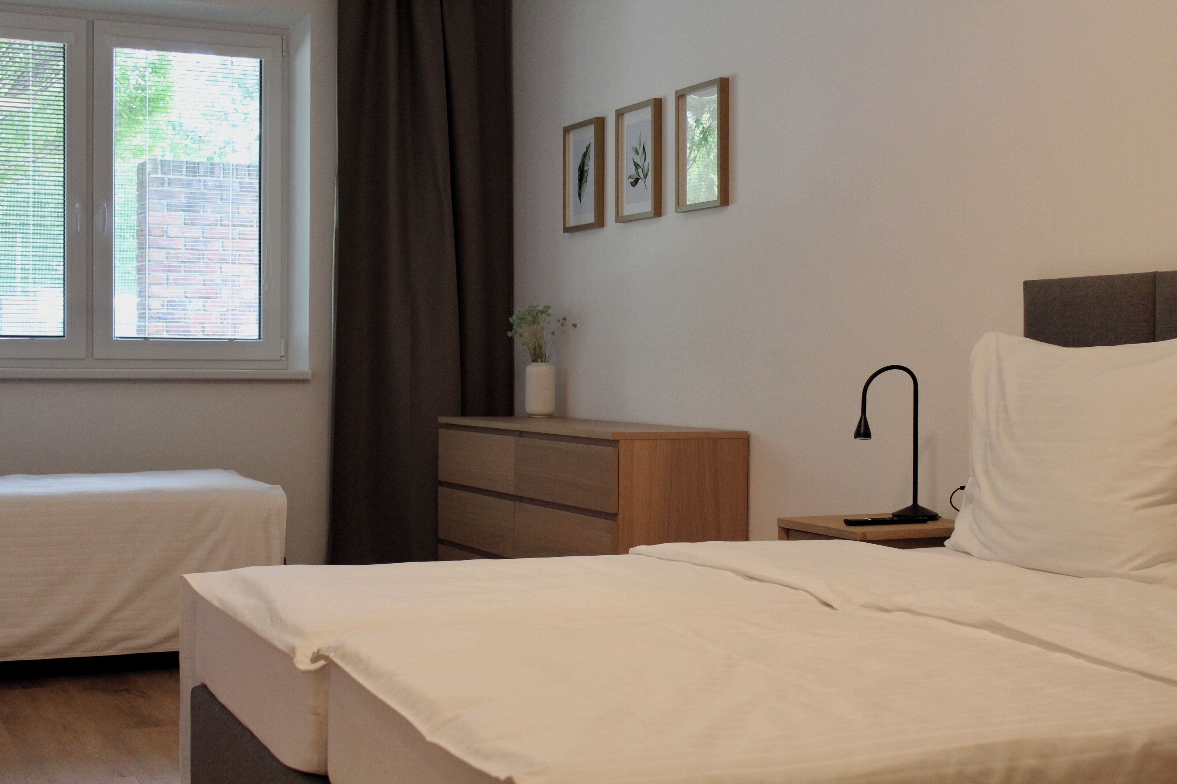 A bedroom with two beds, a nightstand, a lamp, a dresser, a window with blinds, and three framed pictures on the wall.