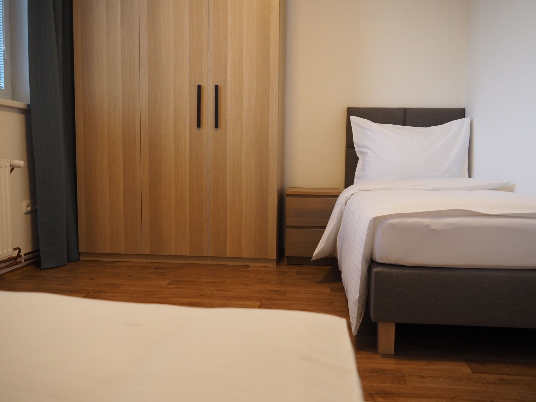 Un dormitorio con un armario de madera, una mesita de noche, una cama con ropa de cama blanca y un cabecero gris.