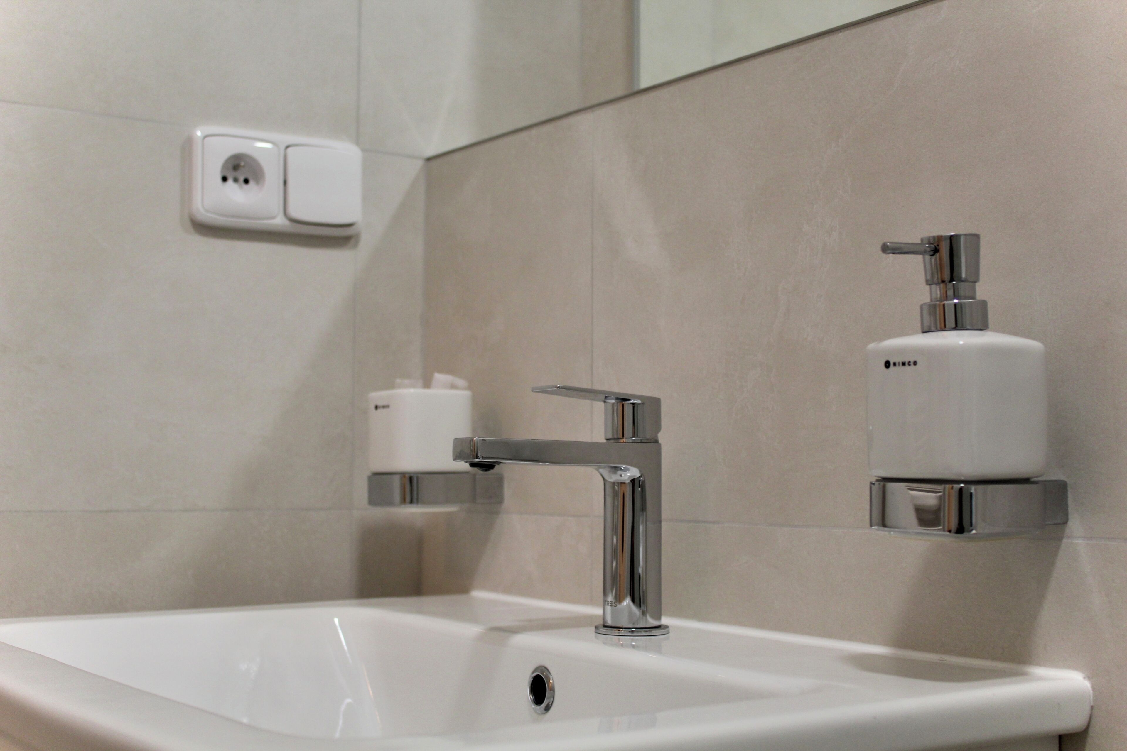 A bathroom with a white sink, chrome faucet, and a soap dispenser mounted on the wall.
