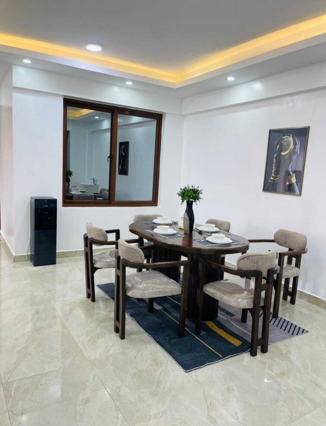 Un comedor con una mesa redonda, seis sillas, un florero con planta, un espejo grande y una obra de arte enmarcada en la pared.