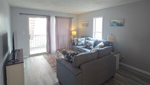 A living room with a grey sofa, a coffee table, and a TV stand. There is a balcony with a view outside. - Cozy Beach Front Retreat (Pensacola Beach)