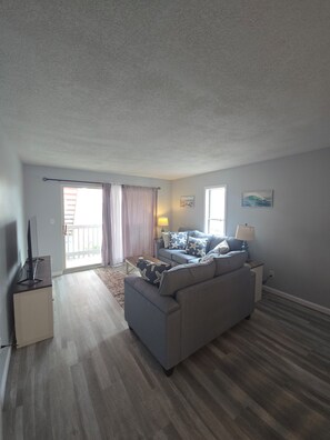 Ein Wohnzimmer mit grauem Sofa, einem Couchtisch und einem TV-Schrank. Es gibt einen Balkon mit Blick ins Freie.