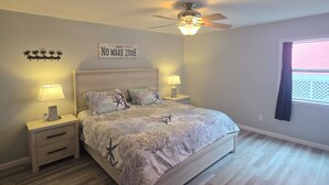 A bedroom with a bed, nightstands, a ceiling fan, a window with curtains, and a wall sign. - Cozy Beach Front Retreat (Pensacola Beach)