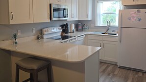 A kitchen with white cabinetry, a central island with a bar stool, a microwave, a stove, and a refrigerator. - Cozy Beach Front Retreat (Pensacola Beach)
