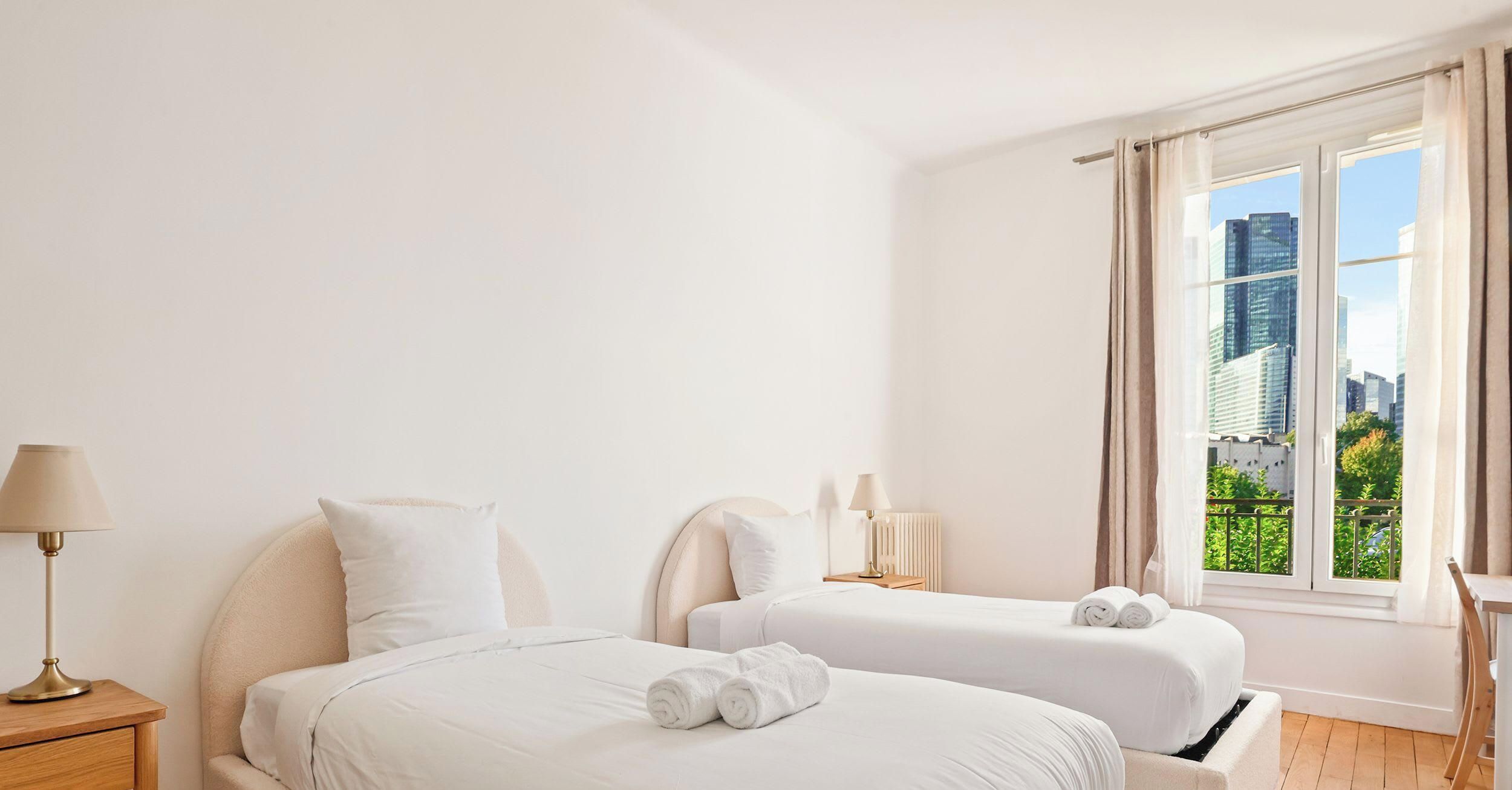 Habitación de hotel con dos camas, una mesita de noche con lámpara, una ventana con cortinas y vistas a la ciudad.