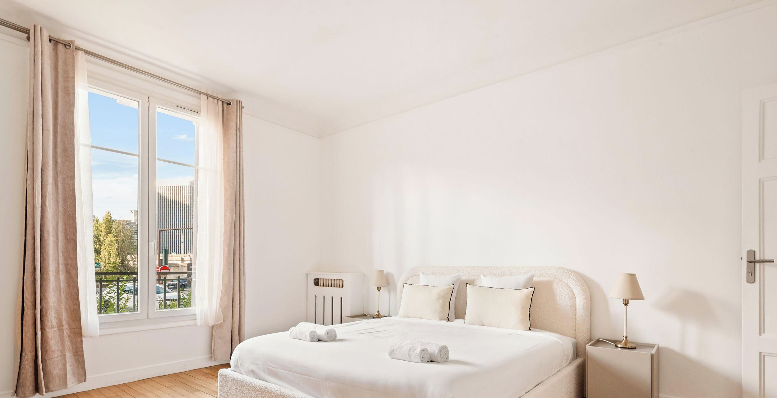 Une chambre d’hôtel avec une grande fenêtre, un lit avec du linge de maison blanc, une table de chevet avec une lampe et un radiateur.