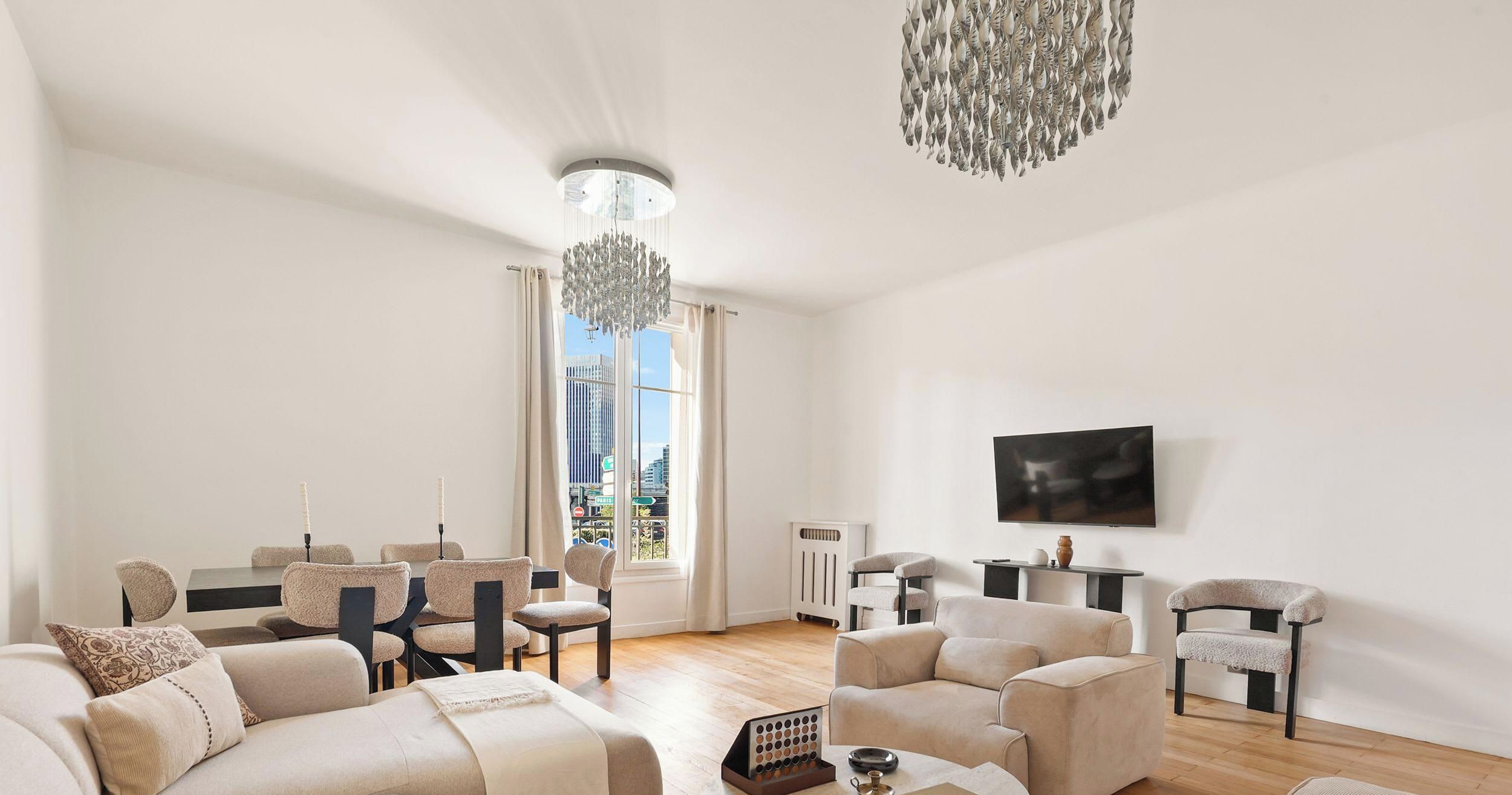 Un vaste salon agrémenté d’un lustre moderne, d’une grande fenêtre offrant une vue sur la ville et d’un téléviseur à écran plat fixé au mur.