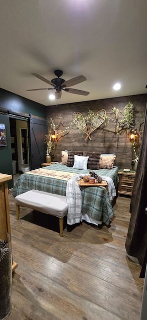 A bedroom with a bed, bench, bedside tables, a ceiling fan, and wall decorations. - Caribou Creek Cabin-Alaska Down South (Hot Springs)
