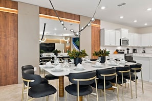 Een moderne eethoek met een witte tafel gedekt voor zes personen, zwarte stoelen met gouden poten, en een keuken op de achtergrond.