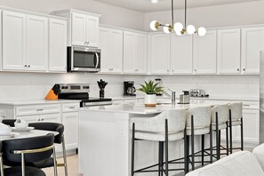 Een moderne keuken met witte kasten, een centraal eiland met barstoelen, een inbouwoven en een hangende lamp.