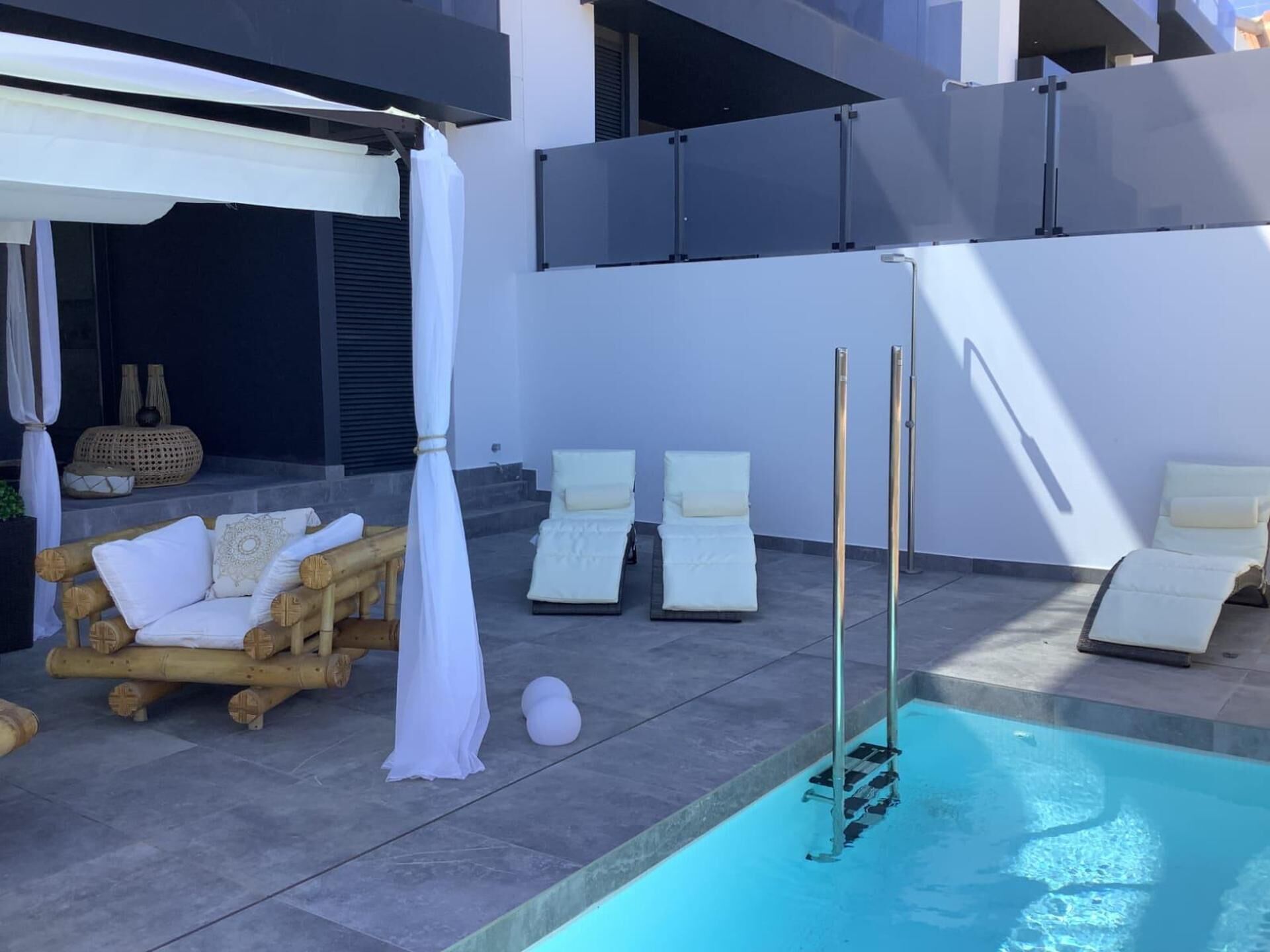 Zona junto a la piscina con sillones de descanso, una banca de madera y una pérgola blanca.
