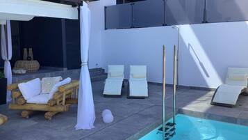 Zona junto a la piscina con sillones de descanso, una banca de madera y una pérgola blanca.