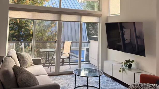 A modern living room with a sofa, a coffee table, a TV, and a balcony with a table and chairs. - The May Townhome - Full Home (Seattle)