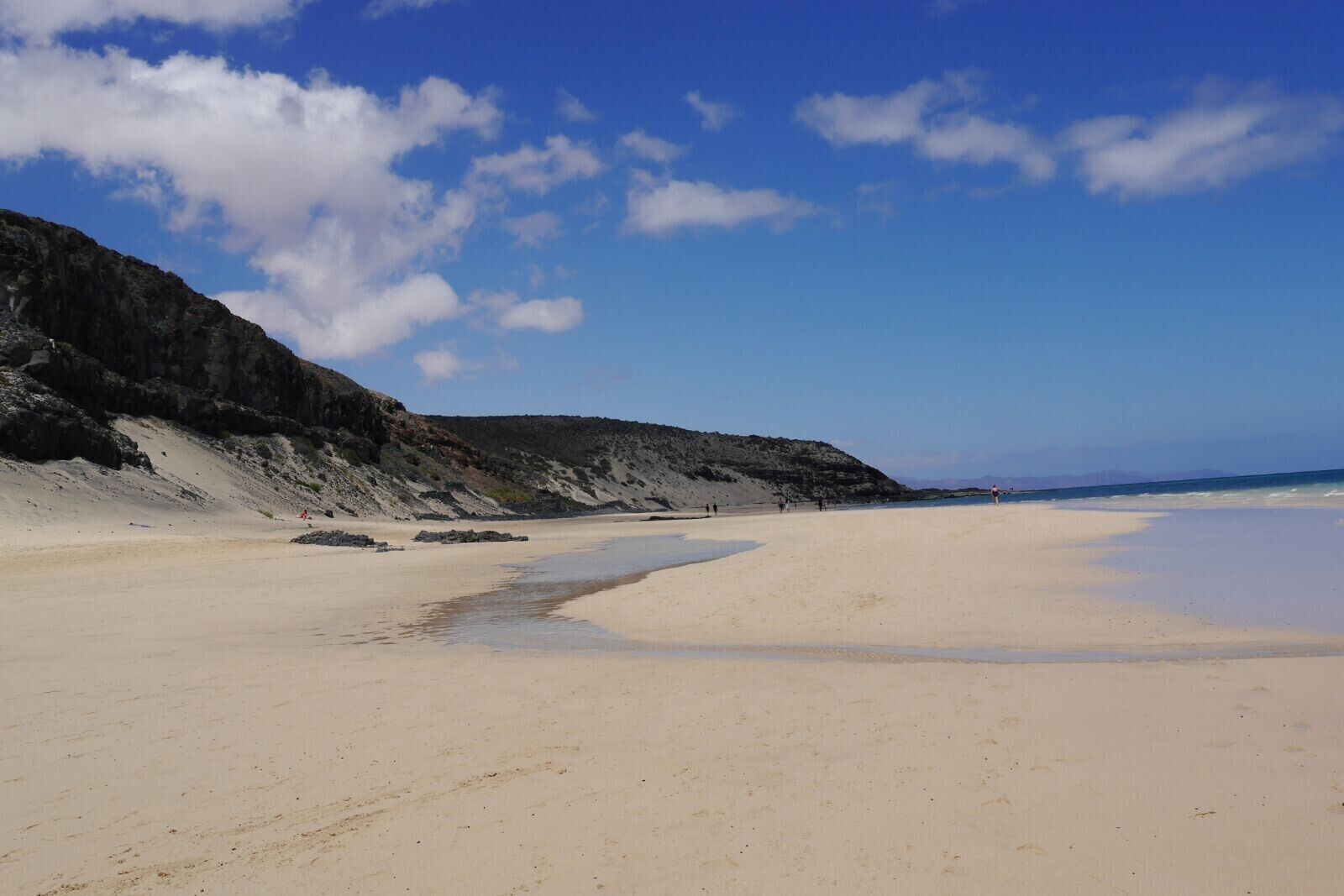 A wide sandy beach with a rocky cliff on one side and a clear blue sky.