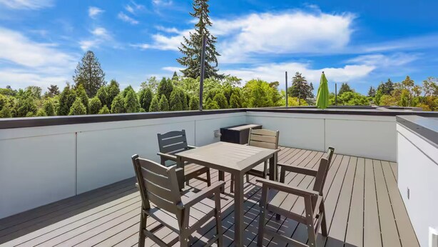 An outdoor deck with a table and chairs, surrounded by a white wall and a view of trees and sky. - Bright & Cozy Seward Park 1BR w/Rooftop Patio + AC (Seattle)