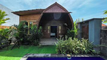 A wooden house with a curved roof, a pool, and lush greenery.
