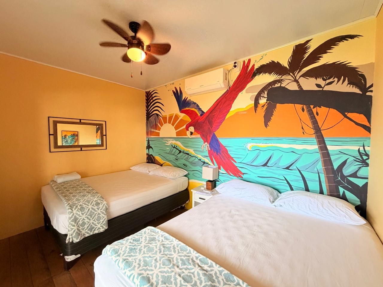 Une chambre d’hôtel avec deux lits, un ventilateur de plafond, un tableau représentant un paysage tropical et un appareil de climatisation.