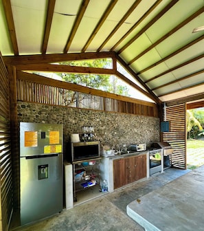 Un área de cocina al aire libre con pared de piedra, vigas de madera, refrigerador, microondas, tostadora y parrilla.