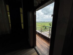 Une pièce dotée d’un plancher en bois et d’une porte donnant sur un balcon avec une rambarde en métal, qui offre une vue sur un champ verdoyant.