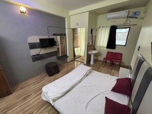 Une chambre d’hôtel avec un lit, un téléviseur fixé au mur, une chaise rouge, un petit tabouret et une fenêtre avec des rideaux.