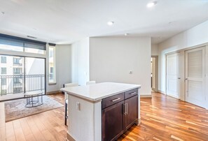 A spacious room with a wooden floor, a kitchen island with dark cabinets, a large window with a balcony, and a door with a louvered design. - Executive 1BR Houston Balcony, Fast Wi-Fi and In-Unit Laundry (Houston)