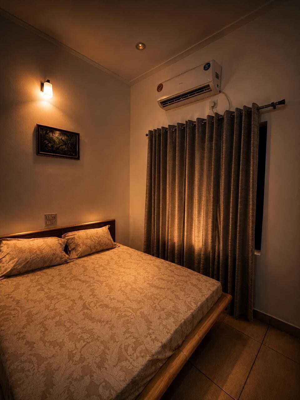 A bedroom with a bed, an air conditioning unit, a wall-mounted light, and a window with curtains.