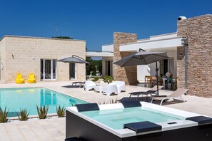A modern outdoor pool area with lounge chairs, umbrellas, and a stone wall. - Trullo Elisa by Perle di Puglia (Carovigno)