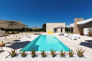A swimming pool with lounge chairs and umbrellas, surrounded by a stone building and greenery. - Trullo Elisa by Perle di Puglia (Carovigno)