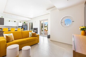 Ein zeitgemäßer Wohnraum mit einem gelben Sofa, weißen Hocker, einem runden Fenster und einer Küchenzeile mit gelben Stühlen.