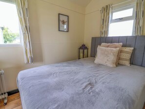 Un dormitorio con cama, un cuadro en la pared, una ventana con cortinas y una mesita de noche con lámpara.