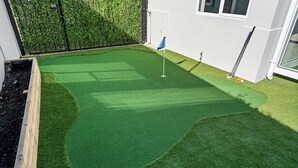 Un’area pratica per il golf su manto erboso sintetico ben curato, con bandiera, circondata da una recinzione in legno e un siepe.