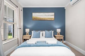 Un dormitorio con una cama, dos mesitas de noche, una lámpara, un cuadro y una ventana con cortinas.