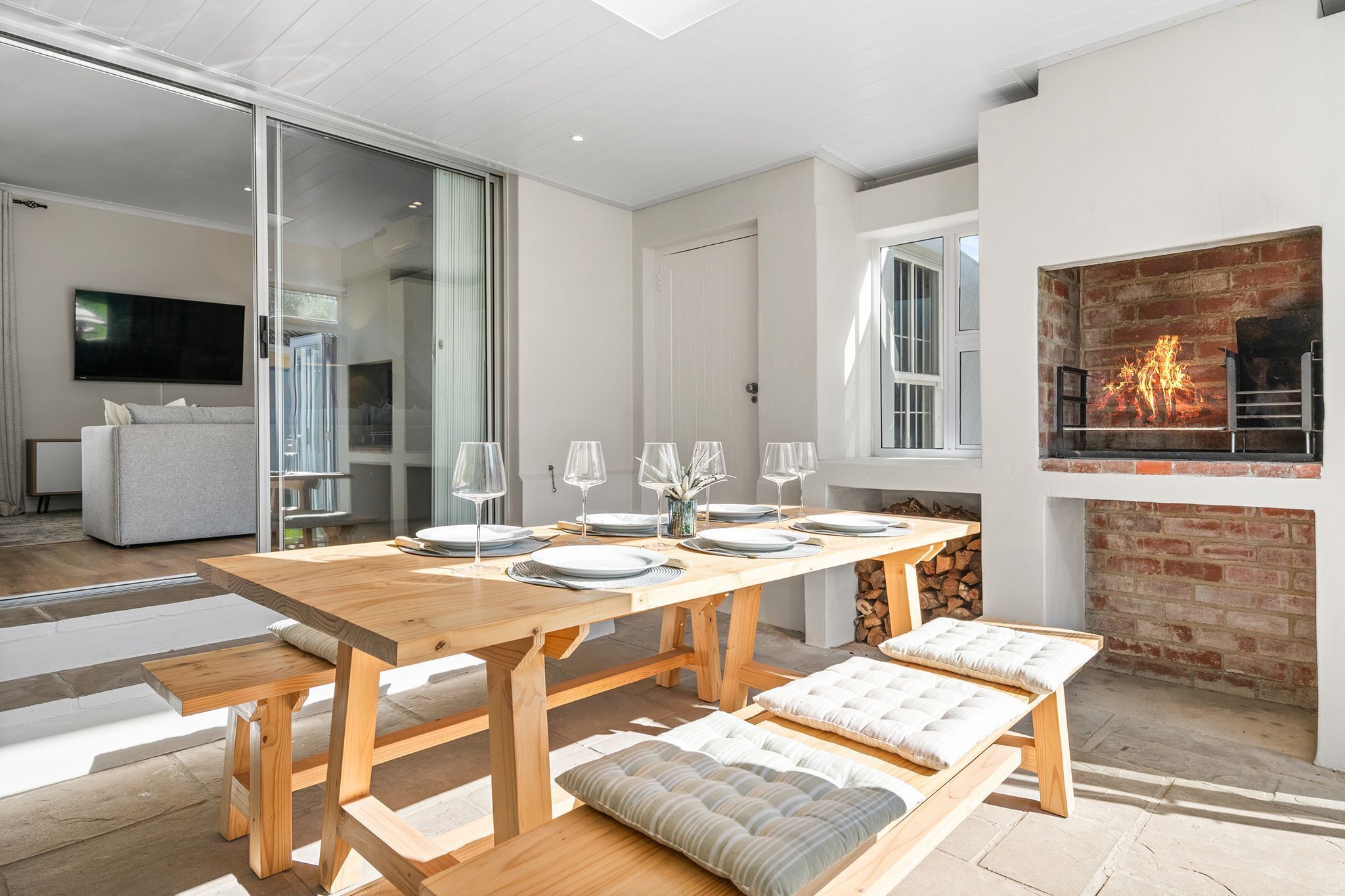Un espace repas moderne avec une table en bois dressée pour quatre personnes, des bancs avec des coussins, une cheminée et un téléviseur en arrière-plan.