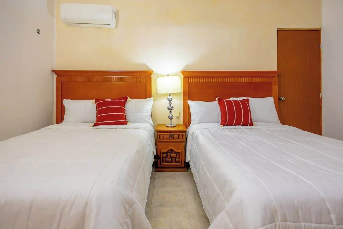 Quarto de hotel com duas camas, uma mesa de cabeceira com abajur e uma porta.