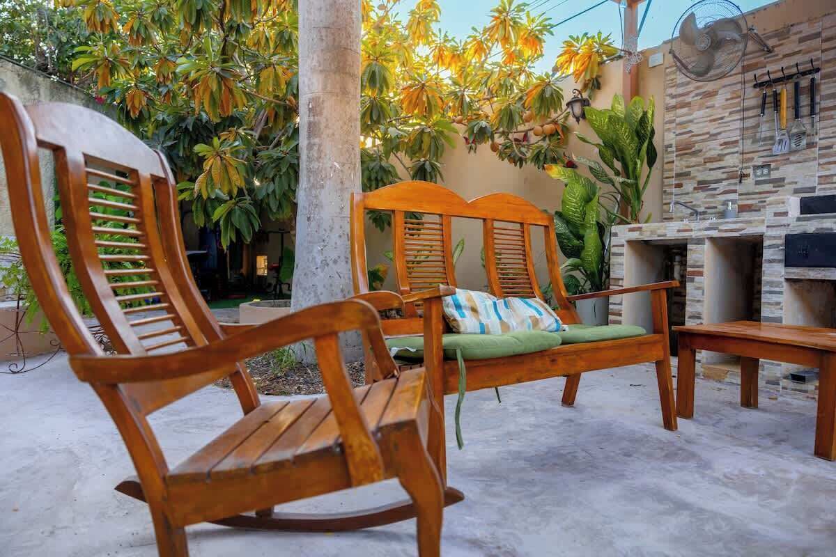 A wooden lounge chair with a cushion, a wooden bench, and a stone wall with a grill.