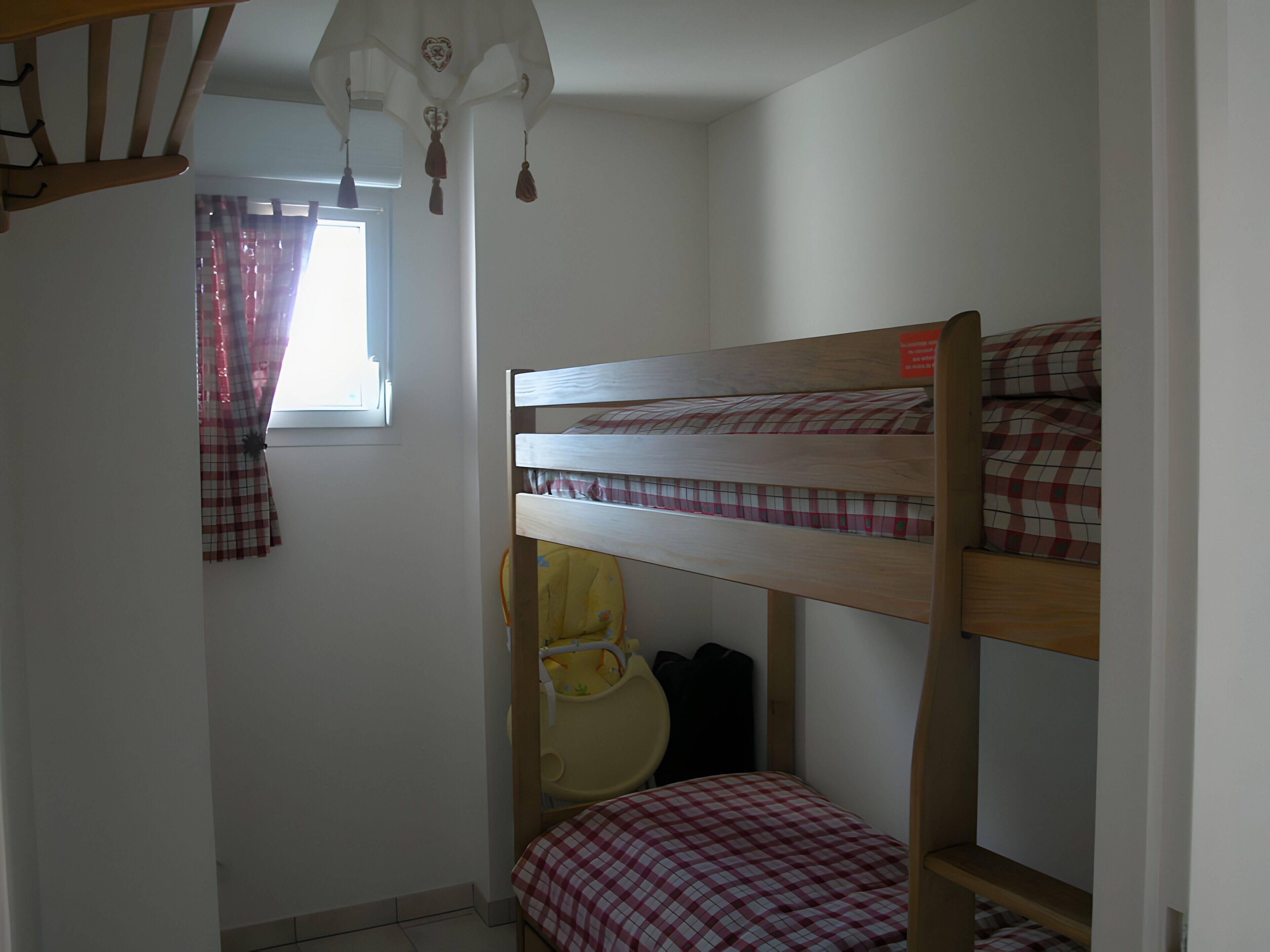 A bunk bed with a checkered bedspread, a small yellow chair, and a window with curtains.