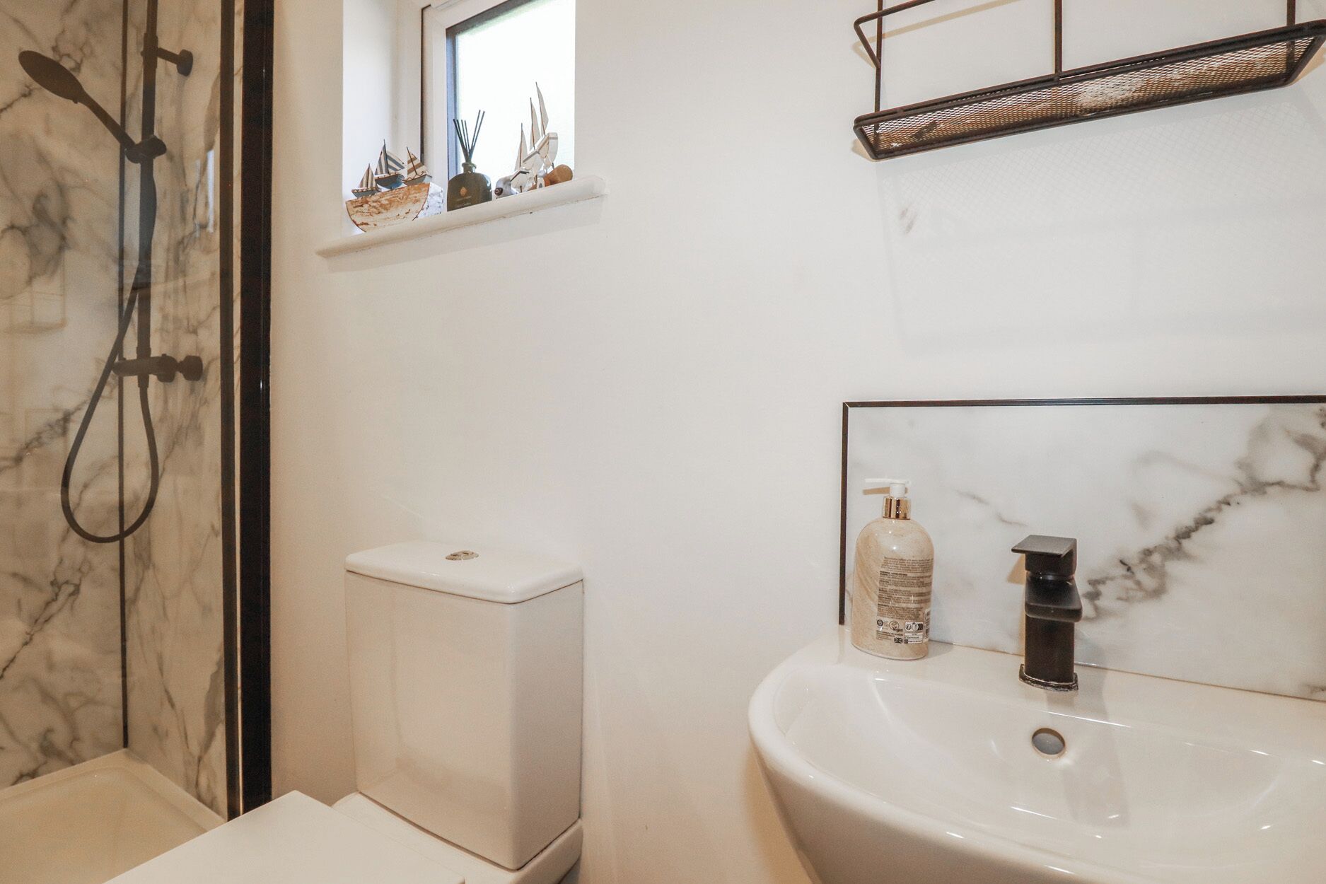 Une salle de bain avec des toilettes blanches, un lavabo doté d’un plan de travail en marbre, une douche avec une porte en verre et une petite fenêtre agrémentée d’objets décoratifs.