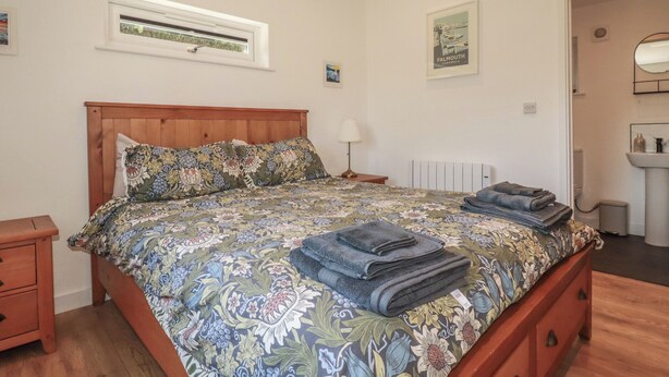 Een slaapkamer met een houten bed, bloemenbedrukking, een nachtkastje, een lamp, een radiator en een ingelijst schilderij, en een badkamer met een wastafel en spiegel.