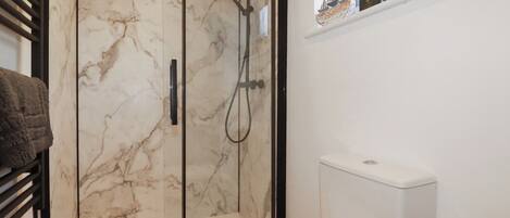 Une salle de bain avec une douche à parois de verre, des toilettes blanches et une petite étagère ornée d’objets décoratifs.