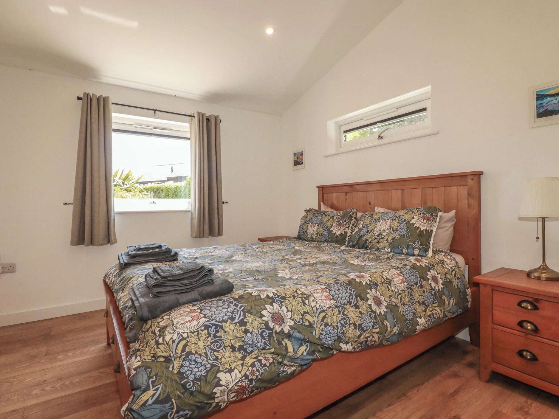 A bedroom with a wooden bed, a nightstand, a window with curtains, and a small table with a lamp.