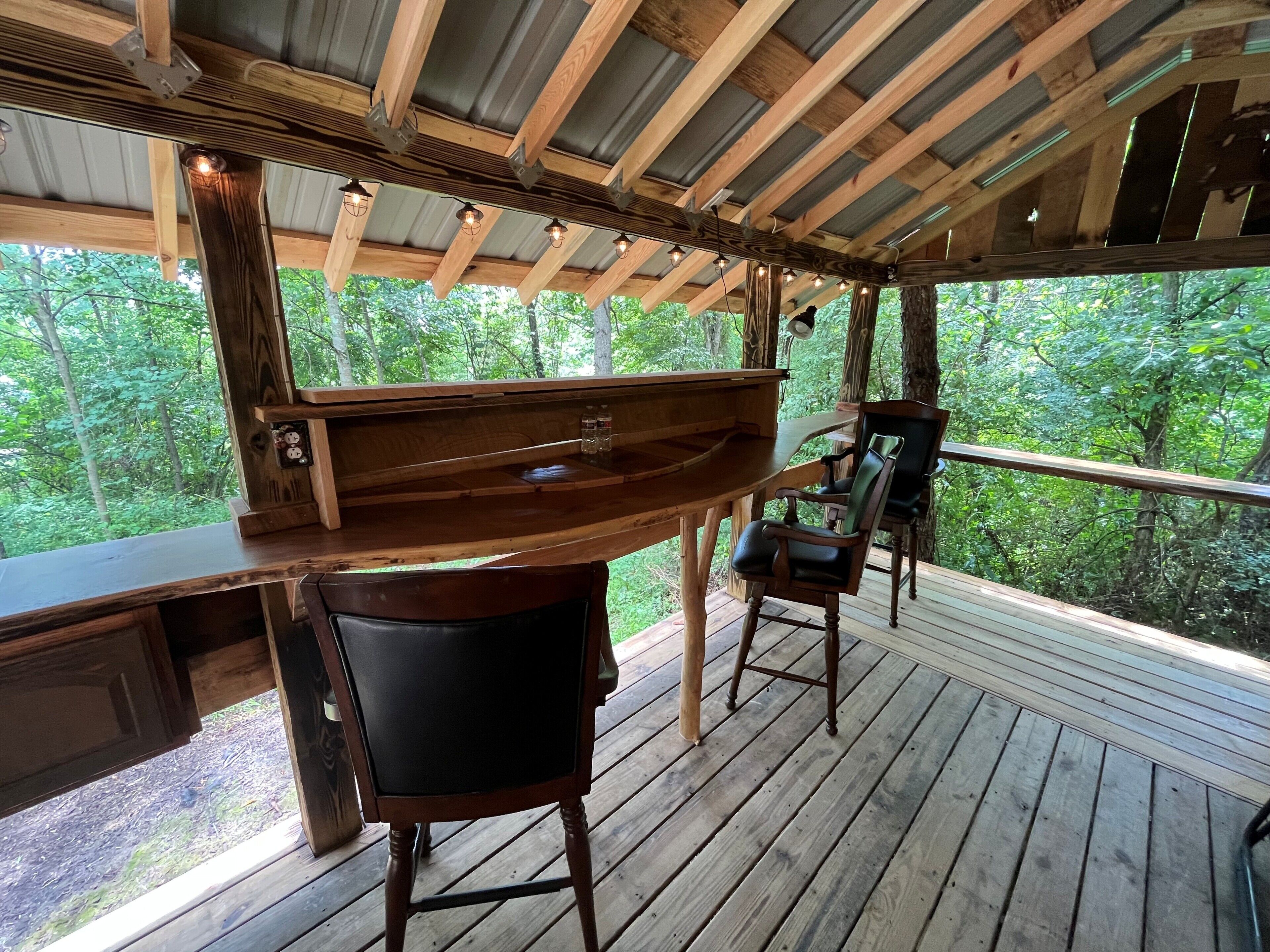 Un muelle de madera con una zona de bar, dos sillas y vista a un bosque.