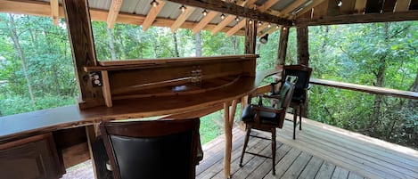 Un muelle de madera con una zona de bar, dos sillas y vista a un bosque.