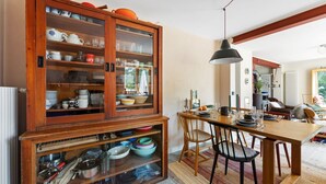 Un coin repas comprenant un buffet en bois contenant de la vaisselle et des ustensiles, une table en bois avec des chaises et un luminaire suspendu.