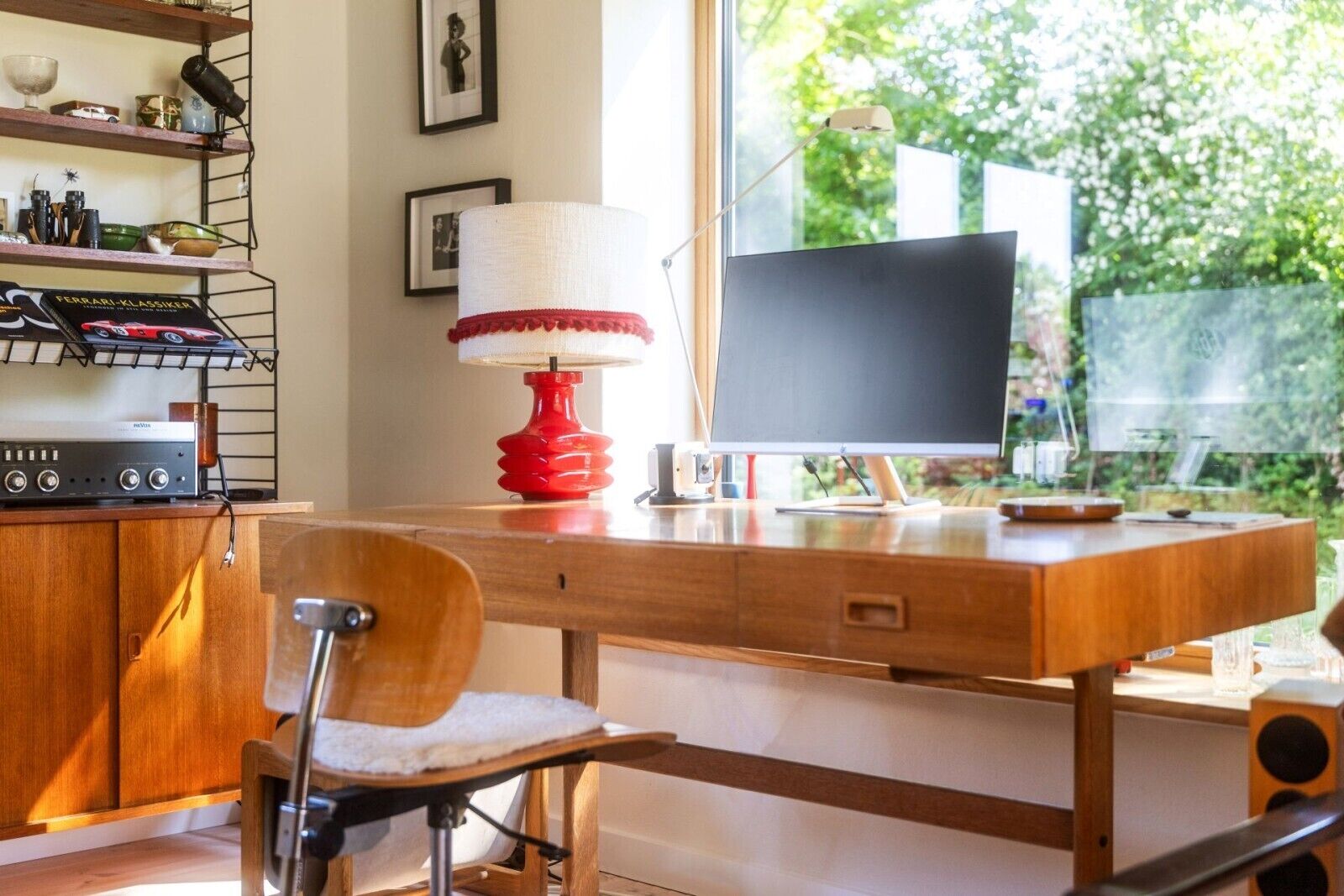 Ein moderner Arbeitsplatz mit einem Holztisch, einer roten Lampe, einem Computermonitor und einem Bücherregal mit verschiedenen Gegenständen.