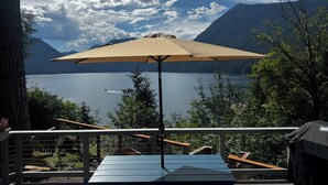 Eine Terrasse mit Tisch und Sonnenschirm mit Blick auf einen See, im Hintergrund Berge.